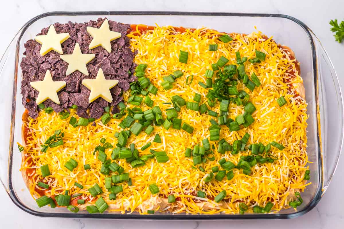A rectangular dish containing a layered dip resembling a flag with cheese stars on top of blue tortilla chips in one corner and shredded cheese with chopped green onions.