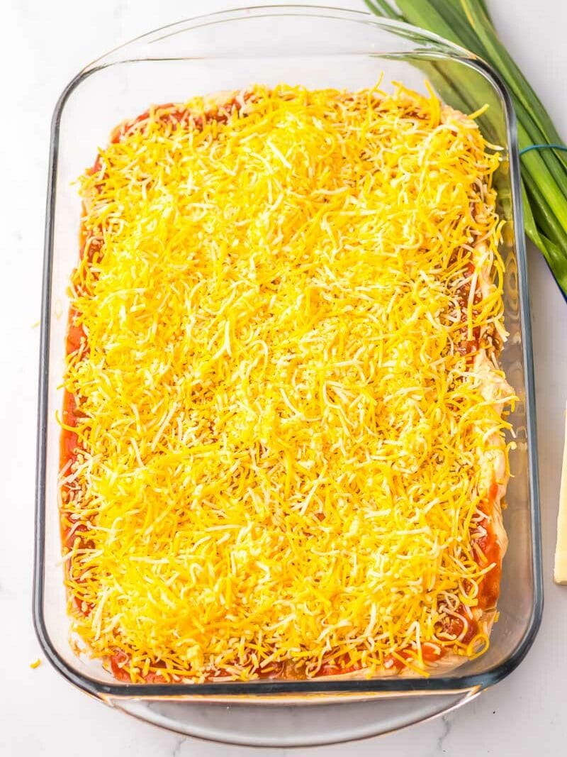 A glass baking dish filled with a layered casserole topped with shredded cheese. Fresh green onions are placed beside the dish on a marble countertop.