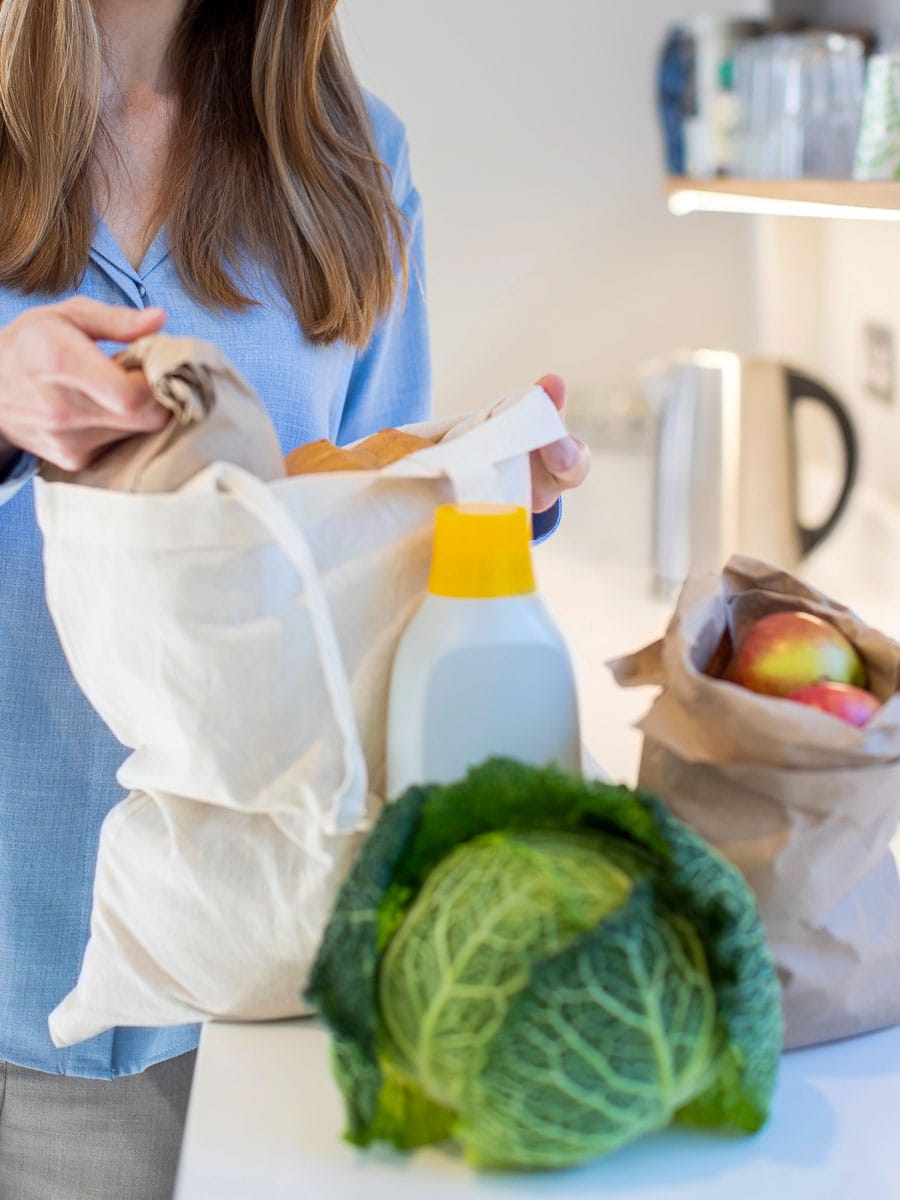 Person in a blue shirt unpacking groceries, including a head of cabbage, a bag of apples, and a bottle of laundry detergent, from reusable bags on a kitchen counter.