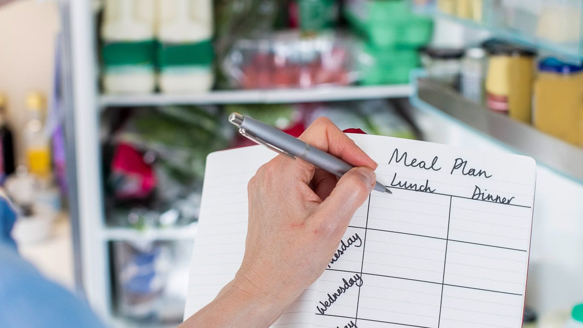 A person writes a meal plan with lunch and dinner columns in a notebook while standing in front of an open refrigerator filled with various food items.