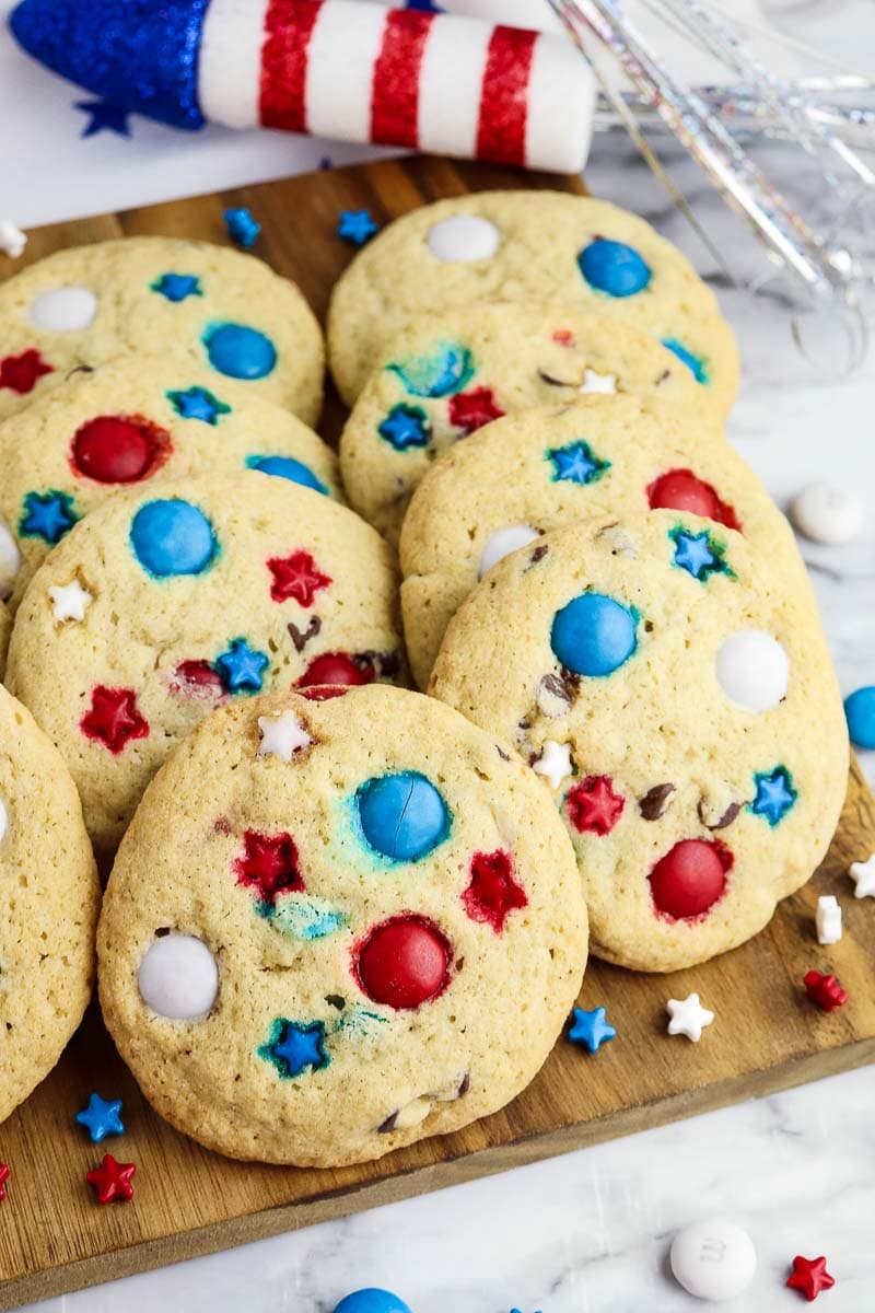 A wooden board with cookies decorated with red, white, and blue candies, some shaped like stars. Fourth of July themed party decorations are in the background.