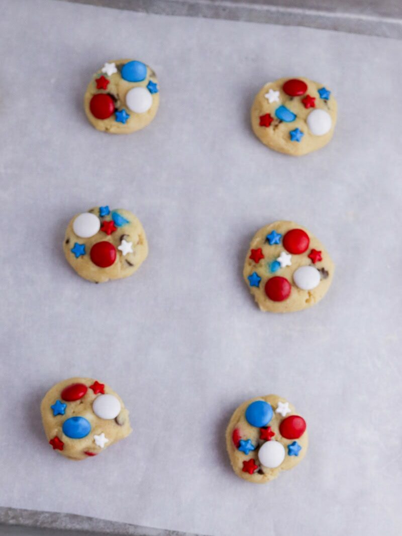 A baking sheet with six round cookies topped with red, white, and blue candies and star-shaped sprinkles. One cookie has a bite taken out of it.