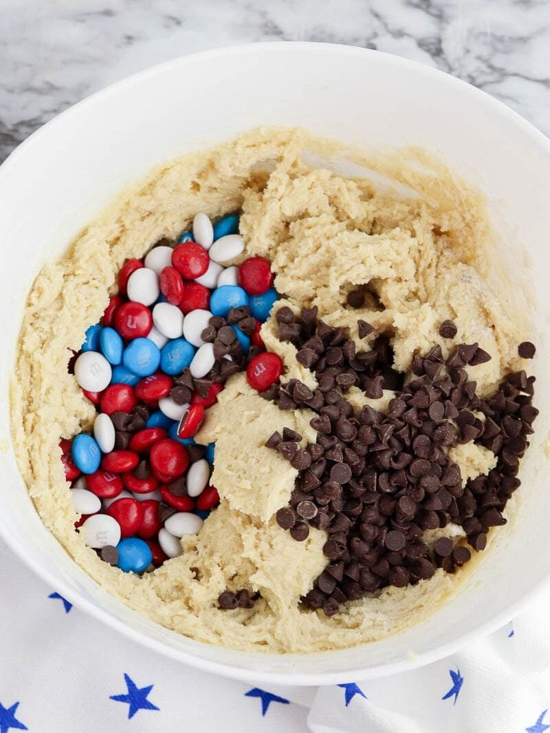A mixing bowl containing cookie dough with red, white, and blue candy-coated chocolates and chocolate chips on top.