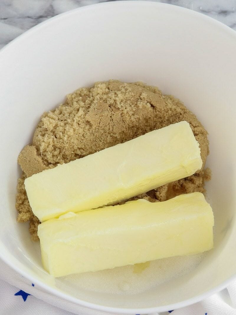 A white bowl containing two sticks of butter and brown sugar on a white cloth with blue stars.