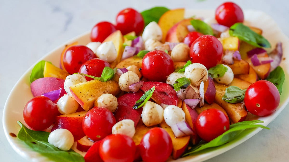A plate of fresh salad with cherry tomatoes, mozzarella balls, peach slices, red onion, and basil leaves, garnished with a drizzle of balsamic vinegar.