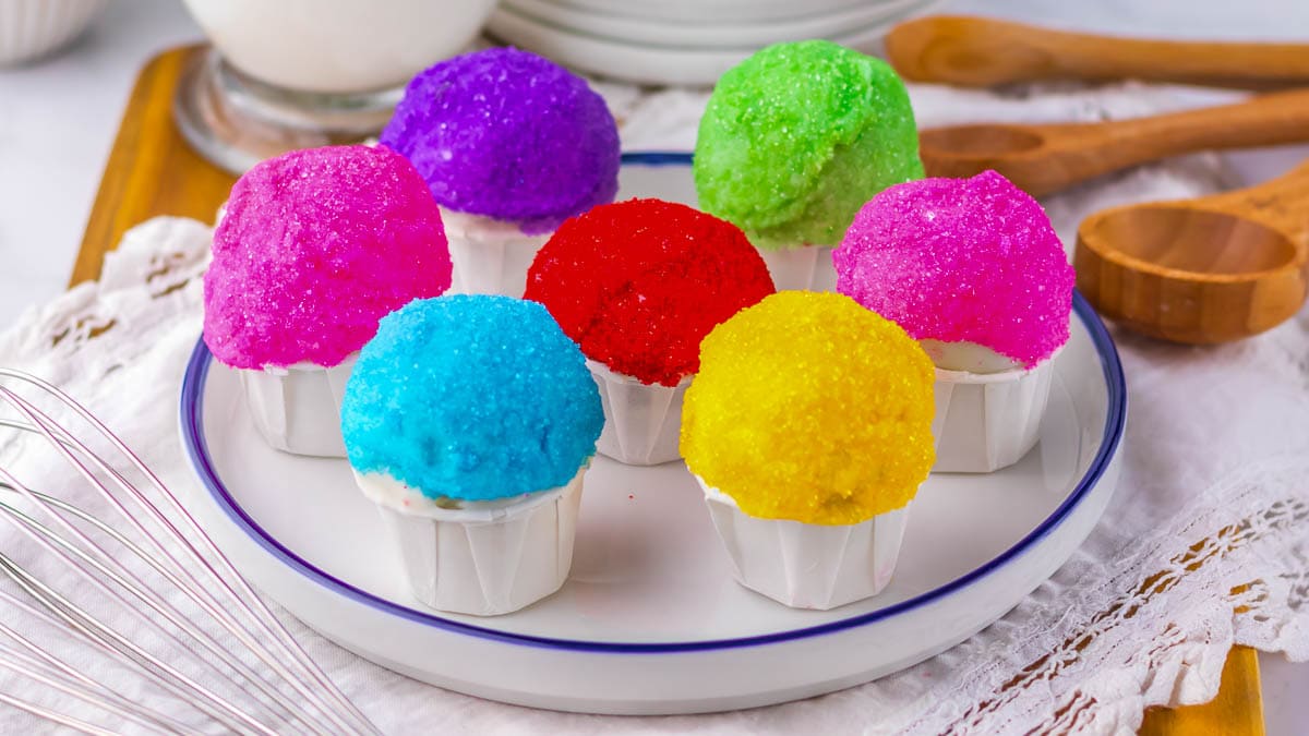 A plate with seven snowcone oreo balls topped with brightly colored sugar crystals sits on a table set with wooden spoons and a whisk.