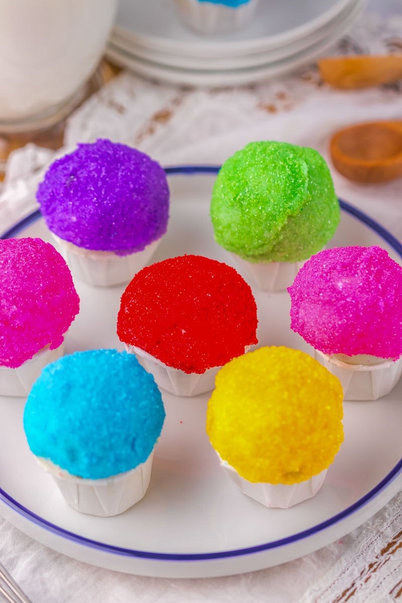 A plate with seven snowcone oreo balls topped with brightly colored sugar crystals sits on a table set with wooden spoons and a whisk.