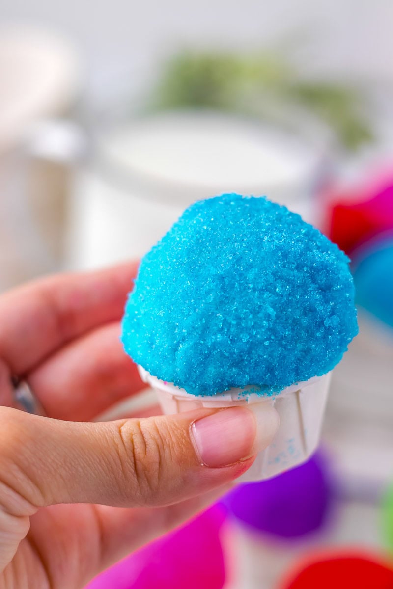 A hand is holding a small cup filled with bright blue snowcone oreo balls. A blurred background features more colored cups and a glass container.